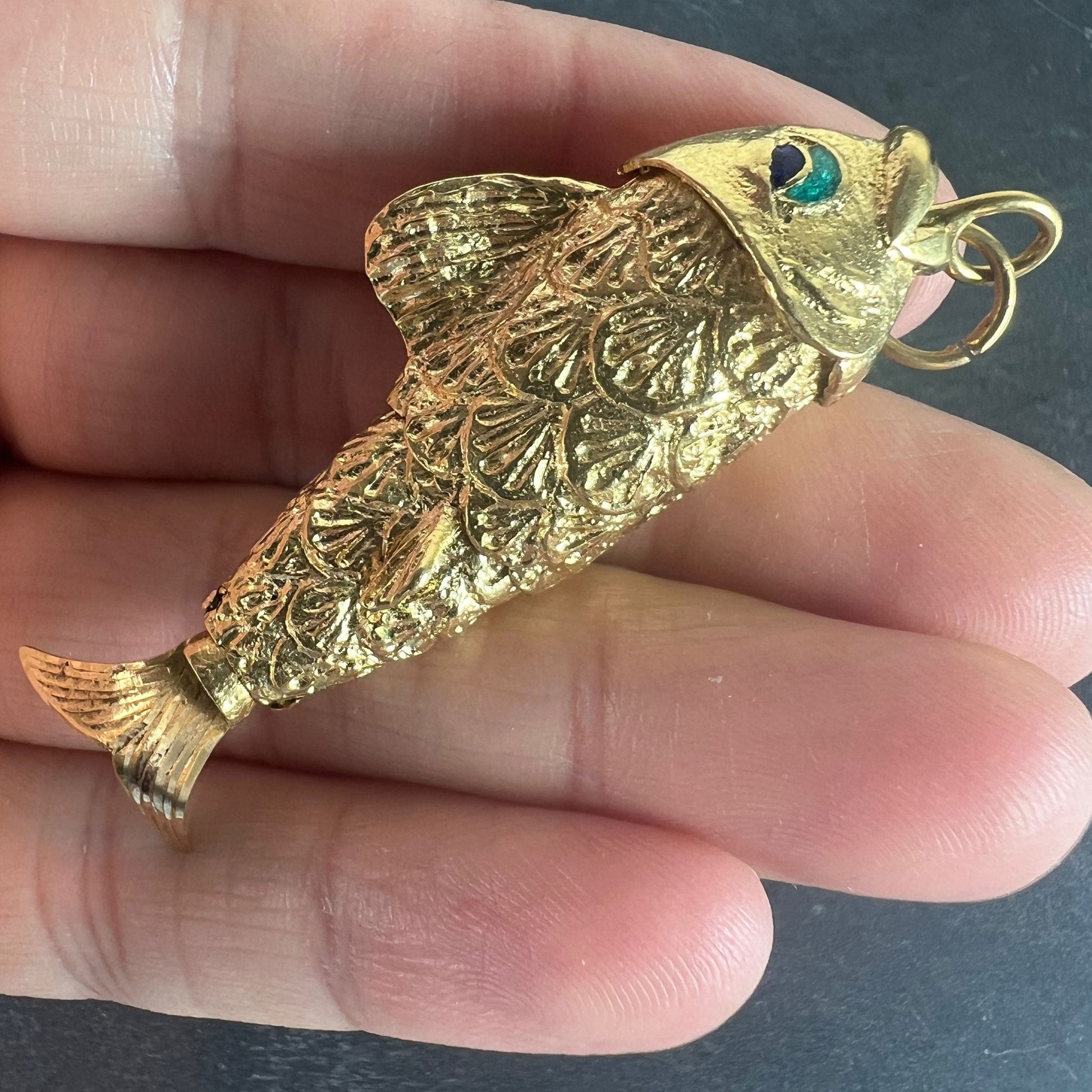A yellow gold fish pendant with a green eye, held in a hand