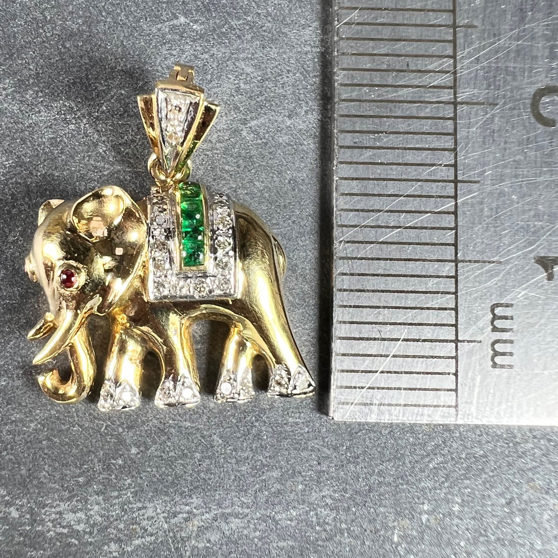 A yellow gold elephant pendant with rubies, emeralds and diamonds, next to a metal ruler on a grey background