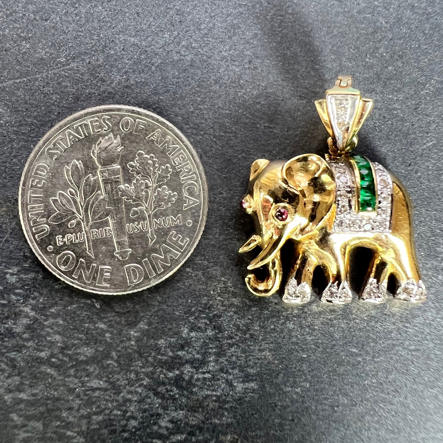 A yellow gold elephant pendant with rubies, emeralds and diamonds, next to a dime coin on a grey background