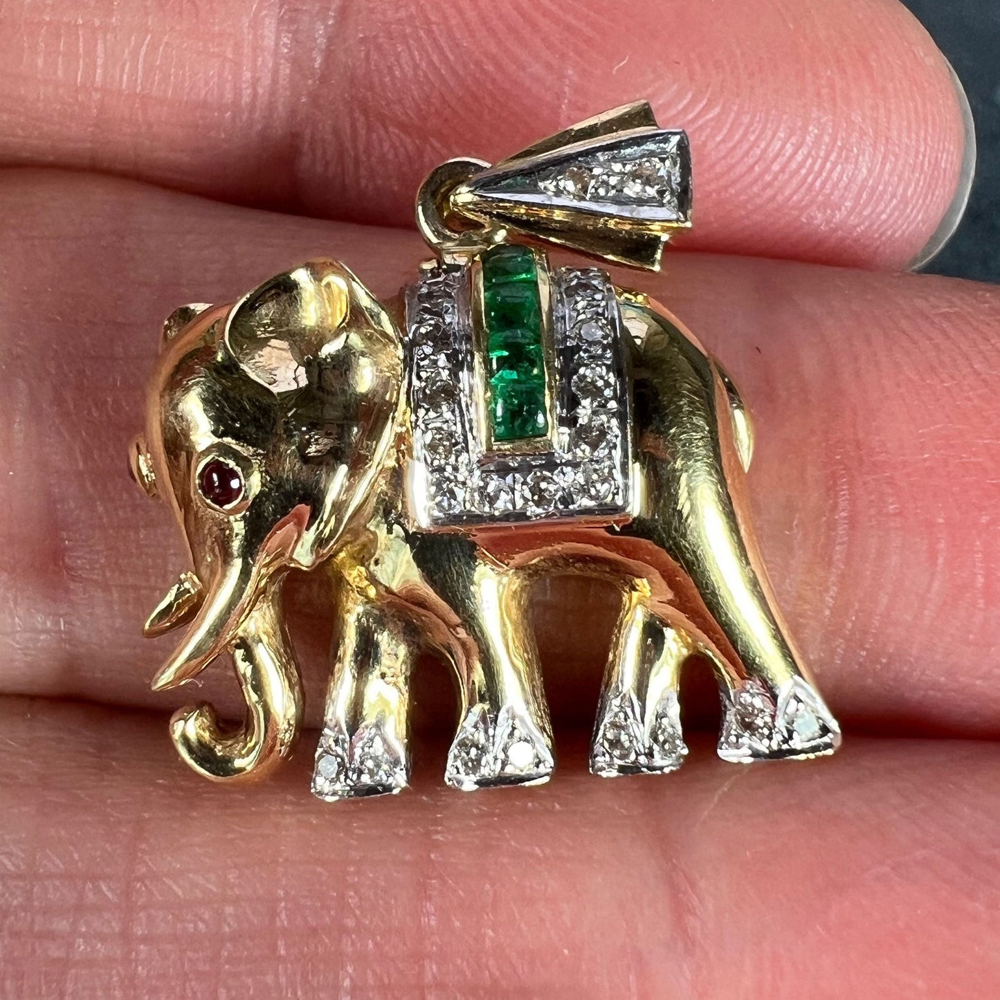 A yellow gold elephant pendant with rubies, emeralds and diamonds, held in a hand on a grey background