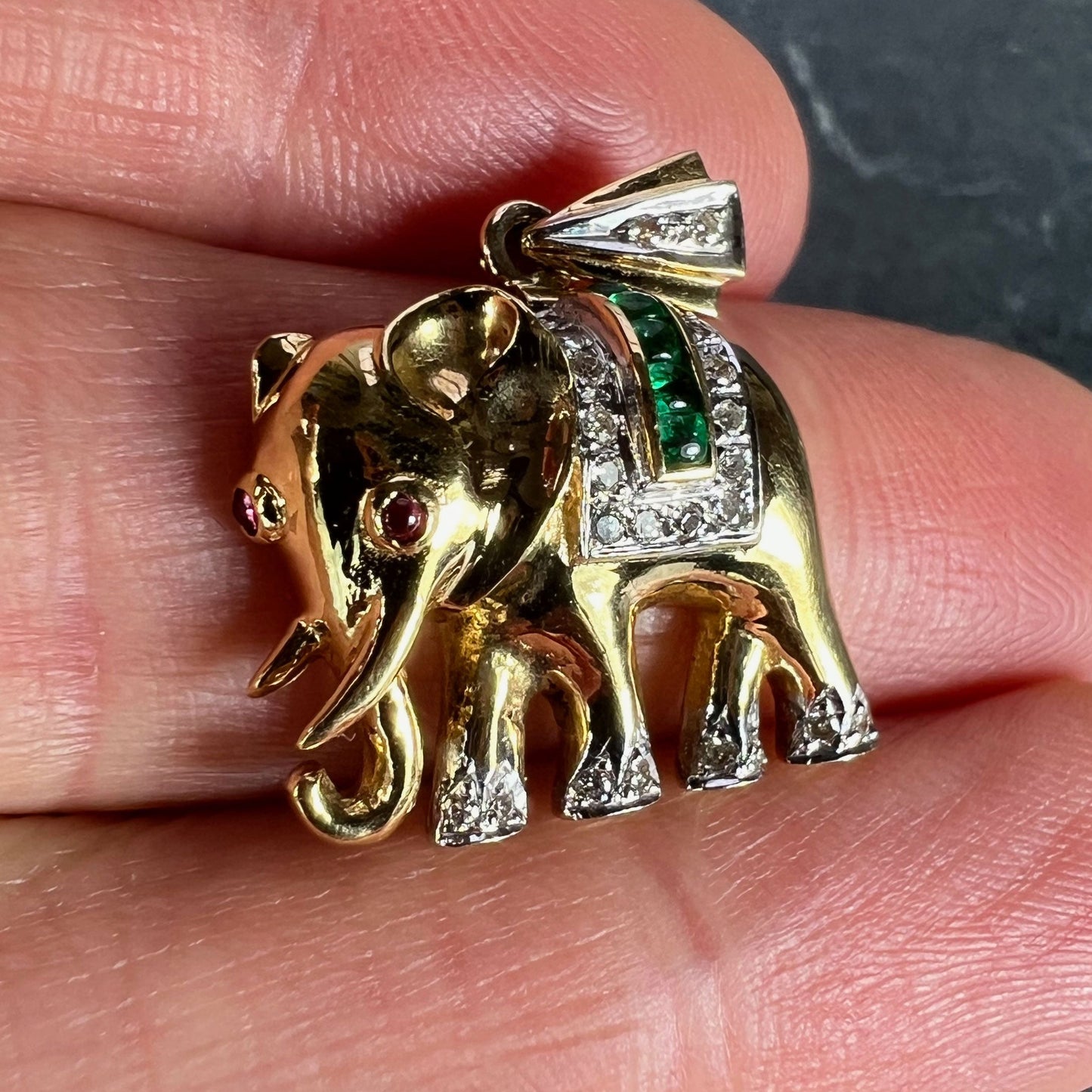 A yellow gold elephant pendant with rubies, emeralds and diamonds, held in a hand on a grey background