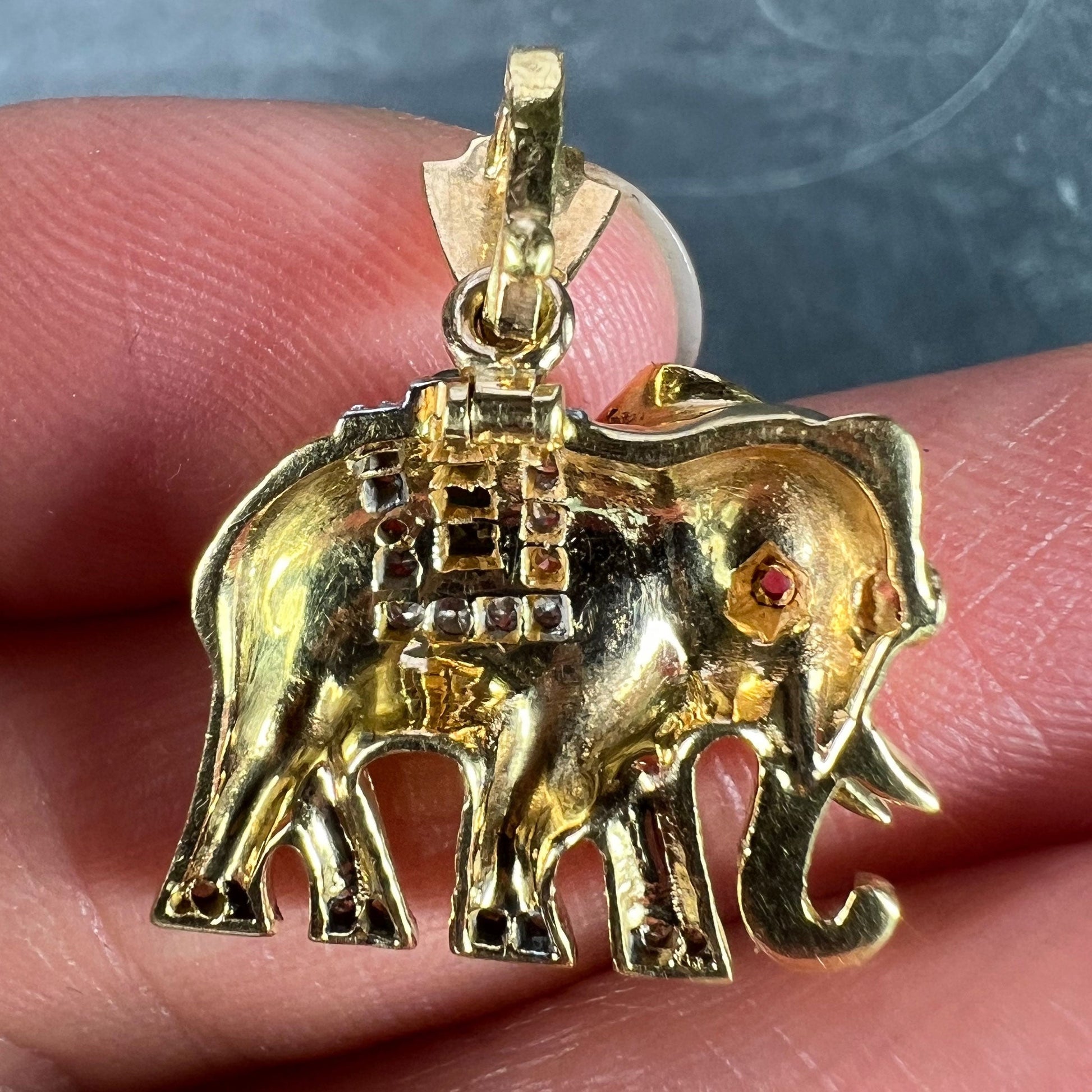 A yellow gold elephant pendant with rubies, emeralds and diamonds, held in a hand on a grey background