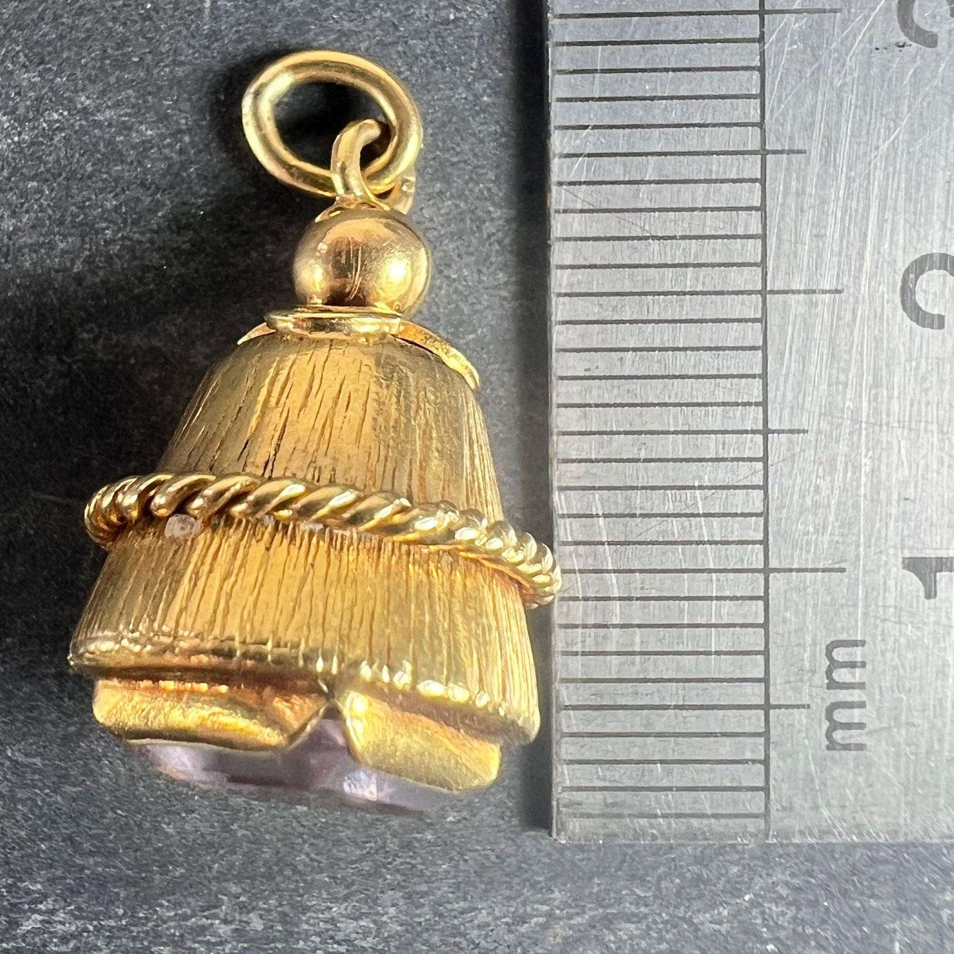 A yellow gold pendant with an amethyst set in the base, next to a metal ruler on a grey background