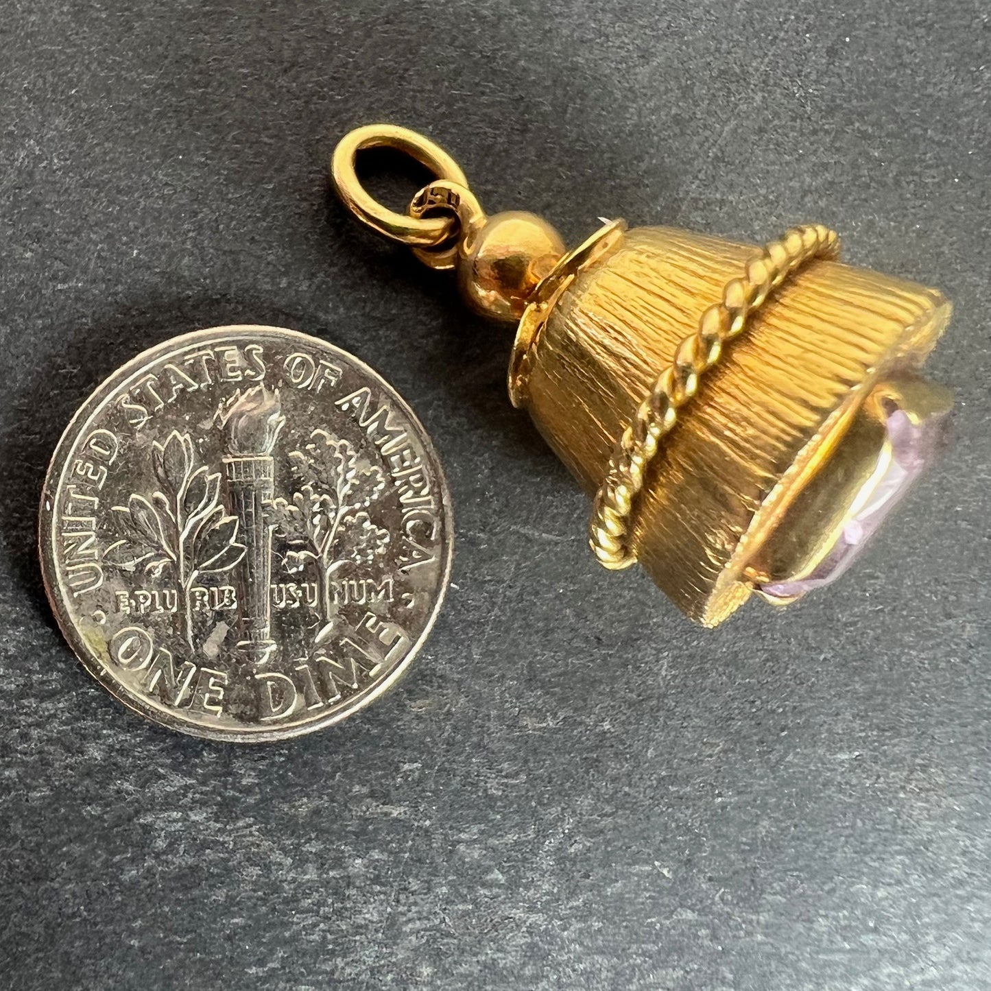 A yellow gold pendant with an amethyst set in the base, next to a dime coin on a grey background