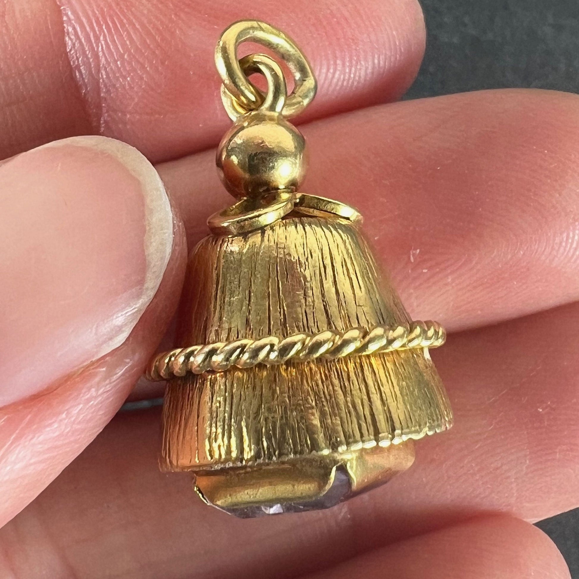 A yellow gold pendant with an amethyst set in the base, held in a hand on a grey background