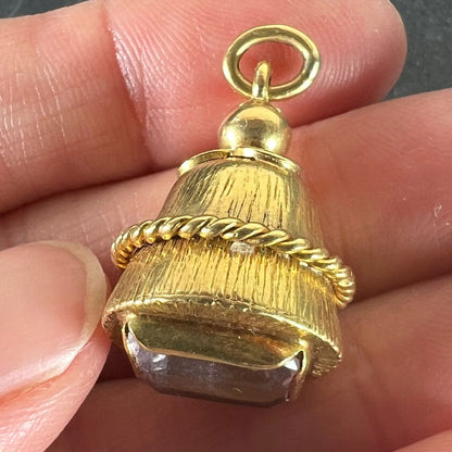 A yellow gold pendant with an amethyst set in the base, held in a hand on a grey background
