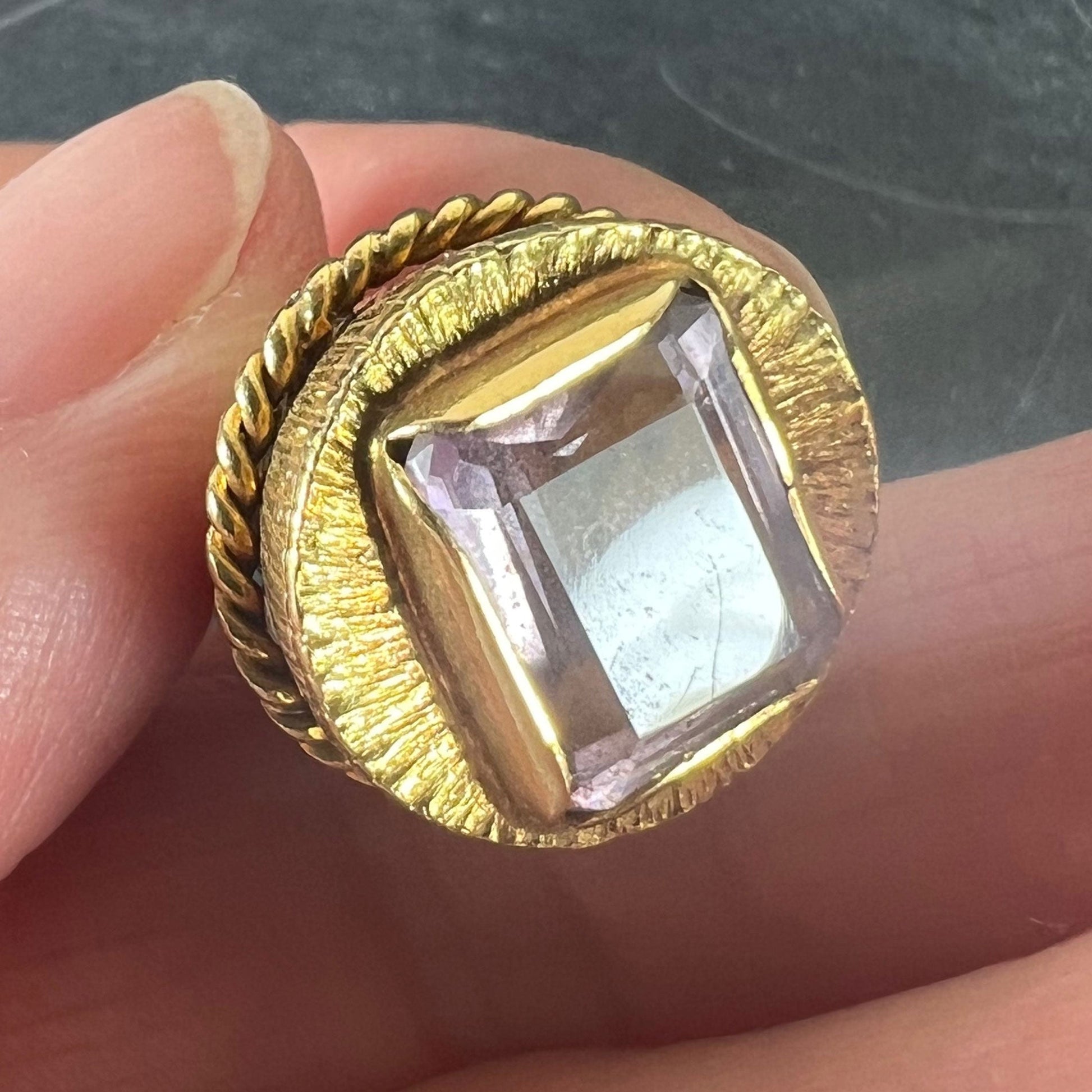 A yellow gold pendant with an amethyst set in the base, held in a hand on a grey background