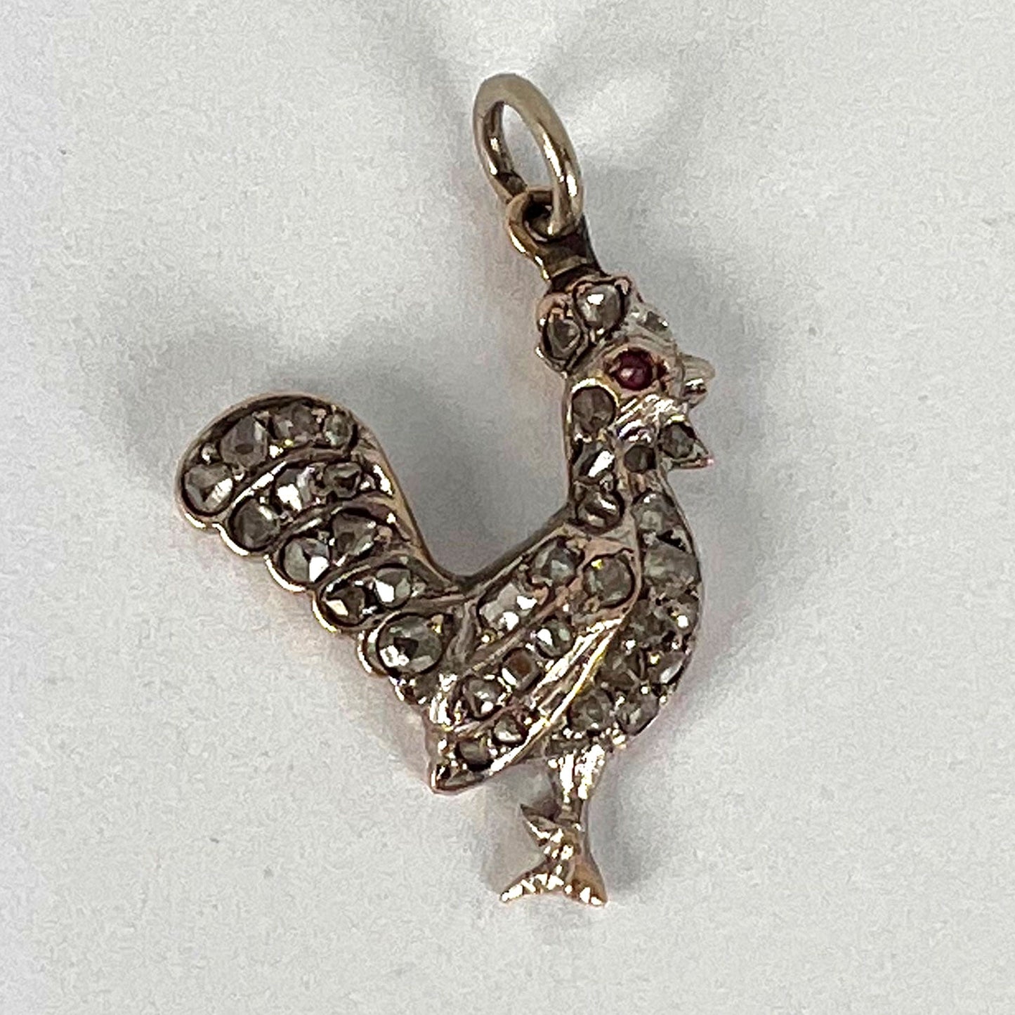 A rose cut diamond rooster pendant with a ruby eye, on a white background