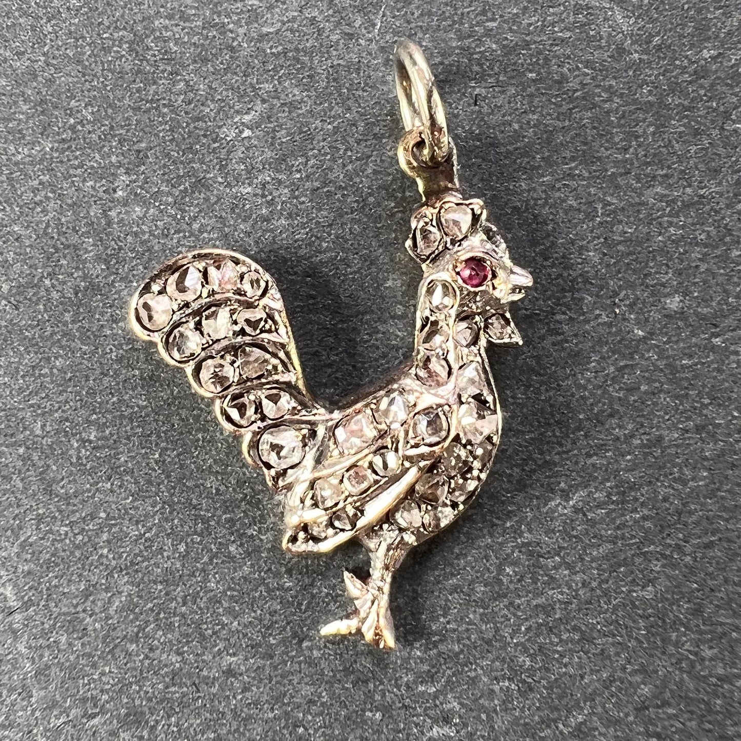 A rose cut diamond rooster pendant with a ruby eye, on a grey background