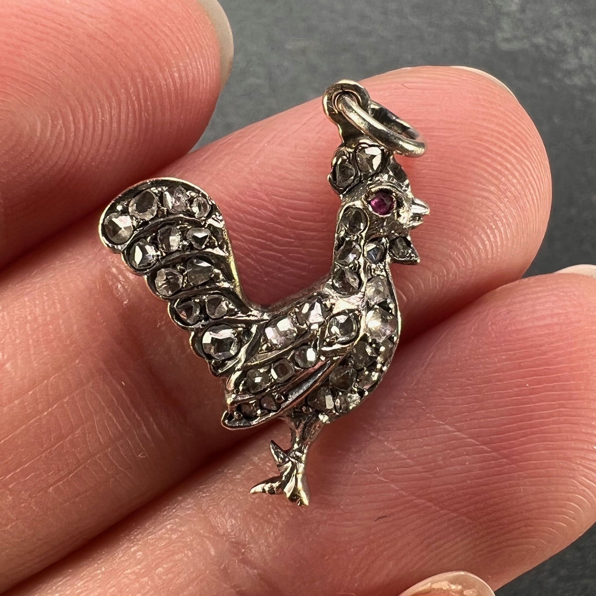 A rose cut diamond rooster pendant with a ruby eye, held in a hand on a grey background