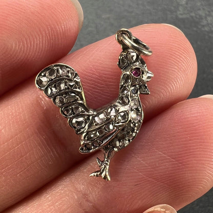 A rose cut diamond rooster pendant with a ruby eye, held in a hand on a grey background