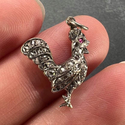 A rose cut diamond rooster pendant with a ruby eye, held in a hand on a grey background