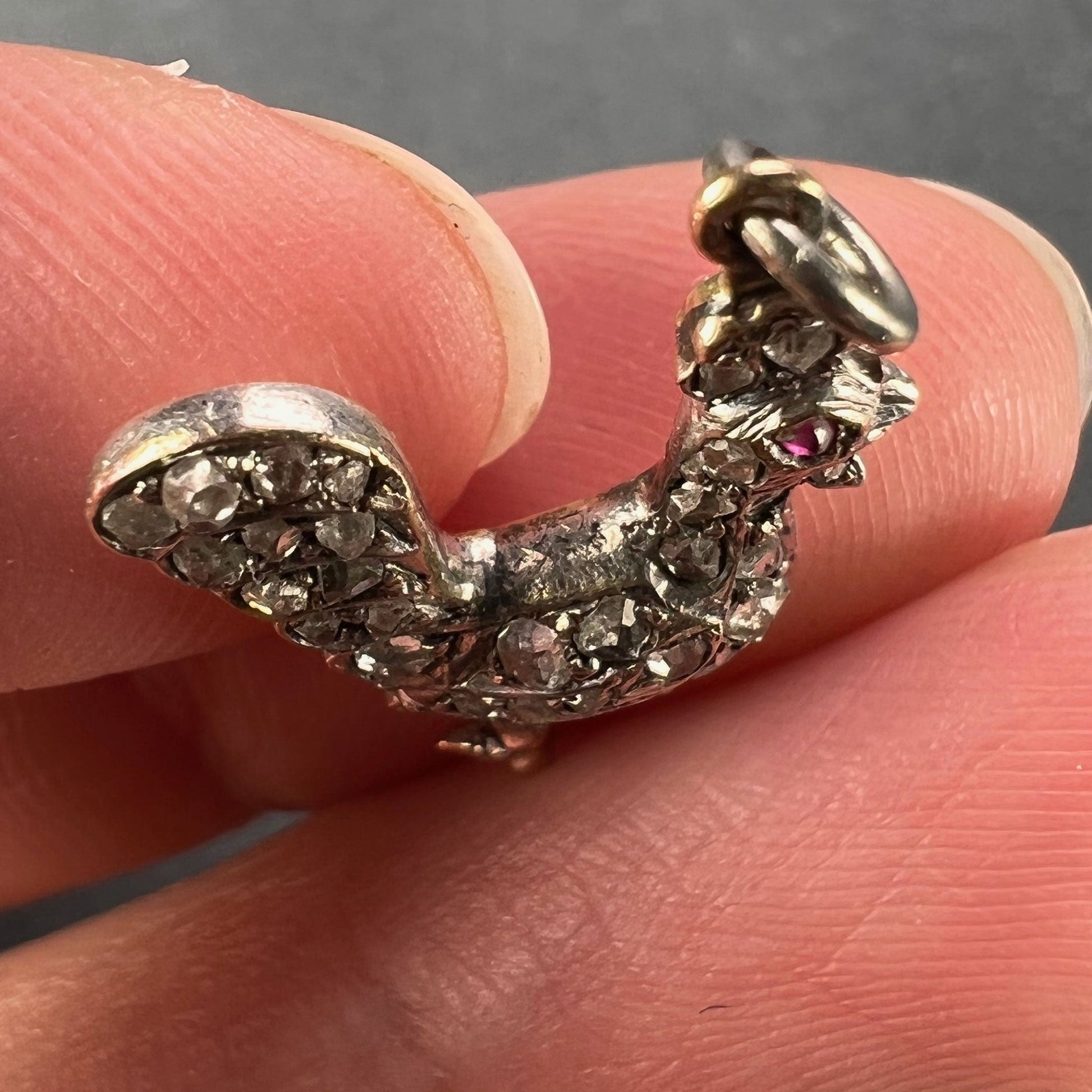 A rose cut diamond rooster pendant with a ruby eye, held in a hand on a grey background
