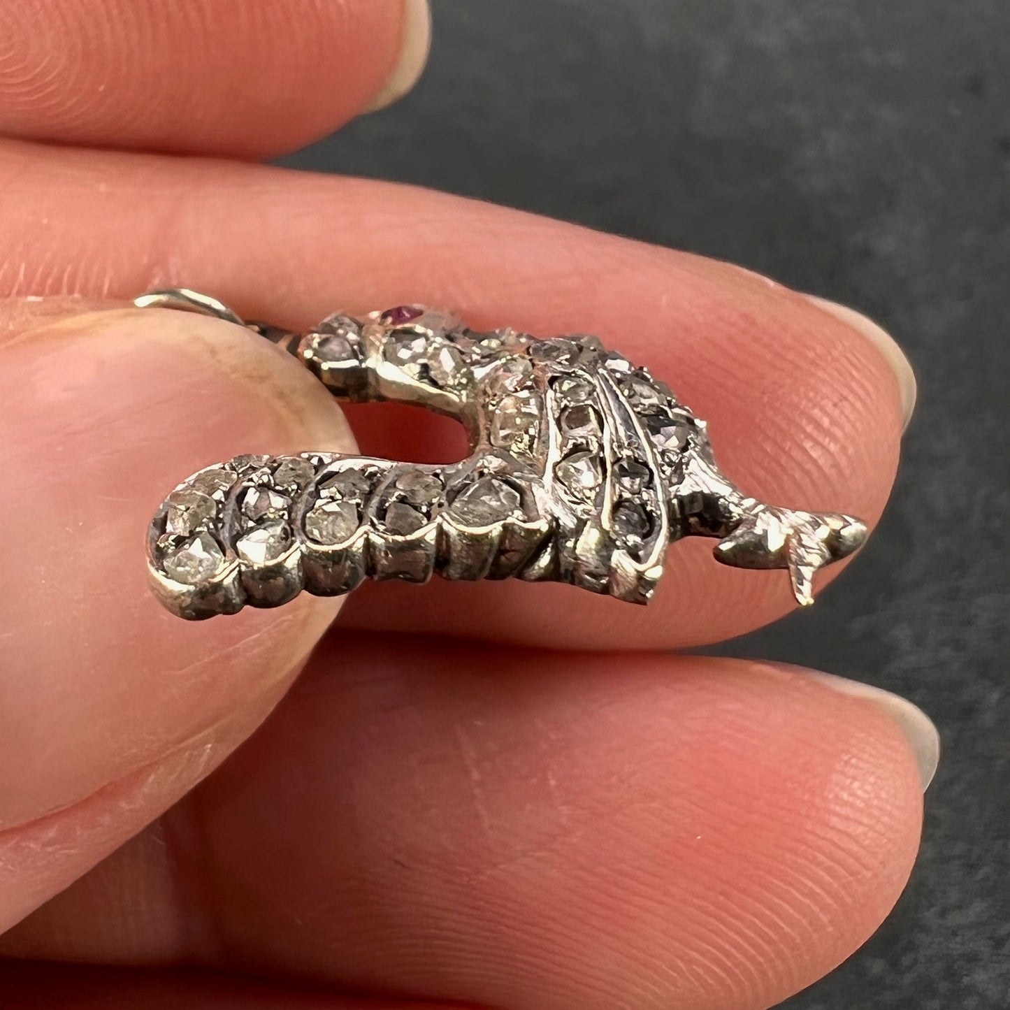 A rose cut diamond rooster pendant with a ruby eye, held in a hand on a grey background