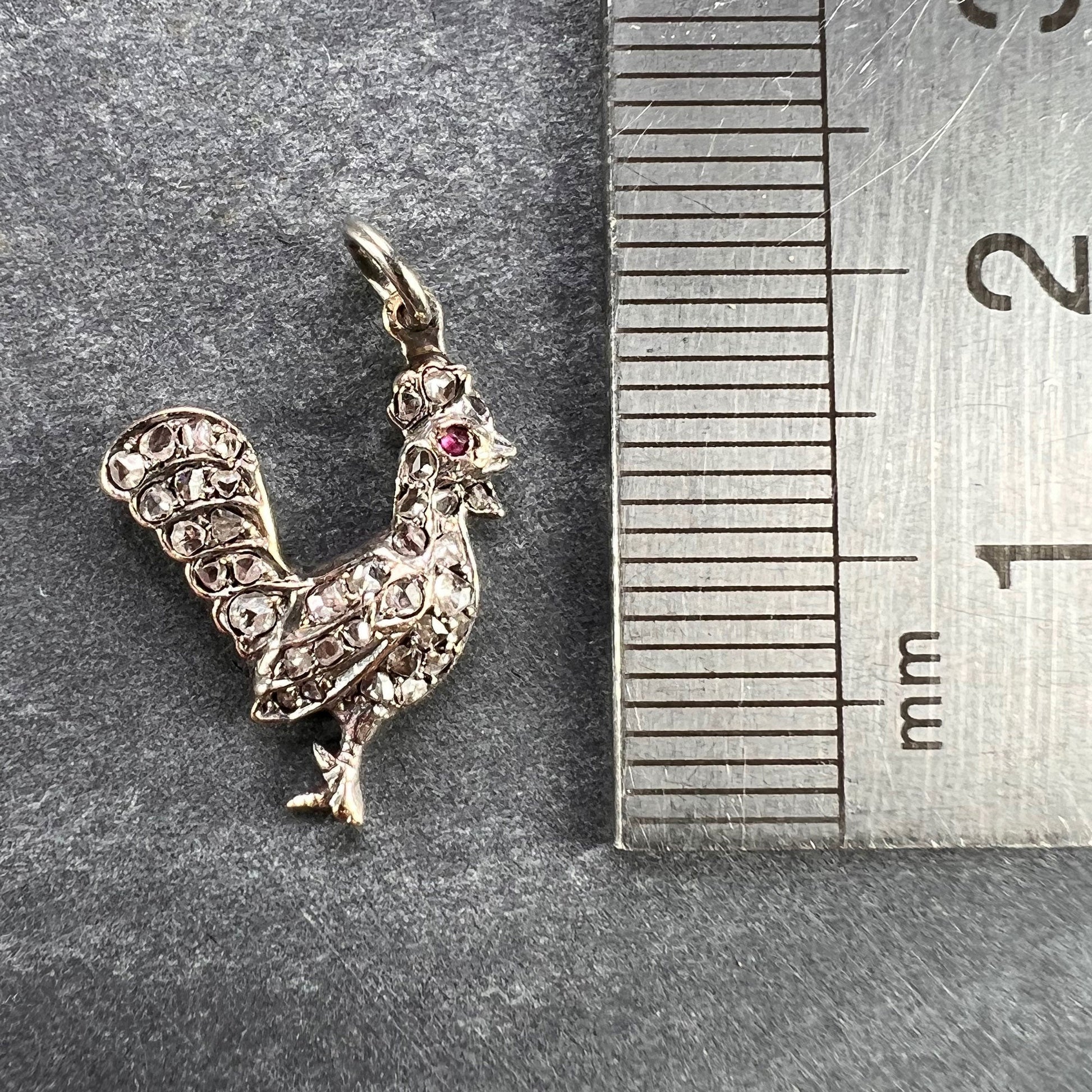 A rose cut diamond rooster pendant with a ruby eye, next to a metal ruler on a grey background