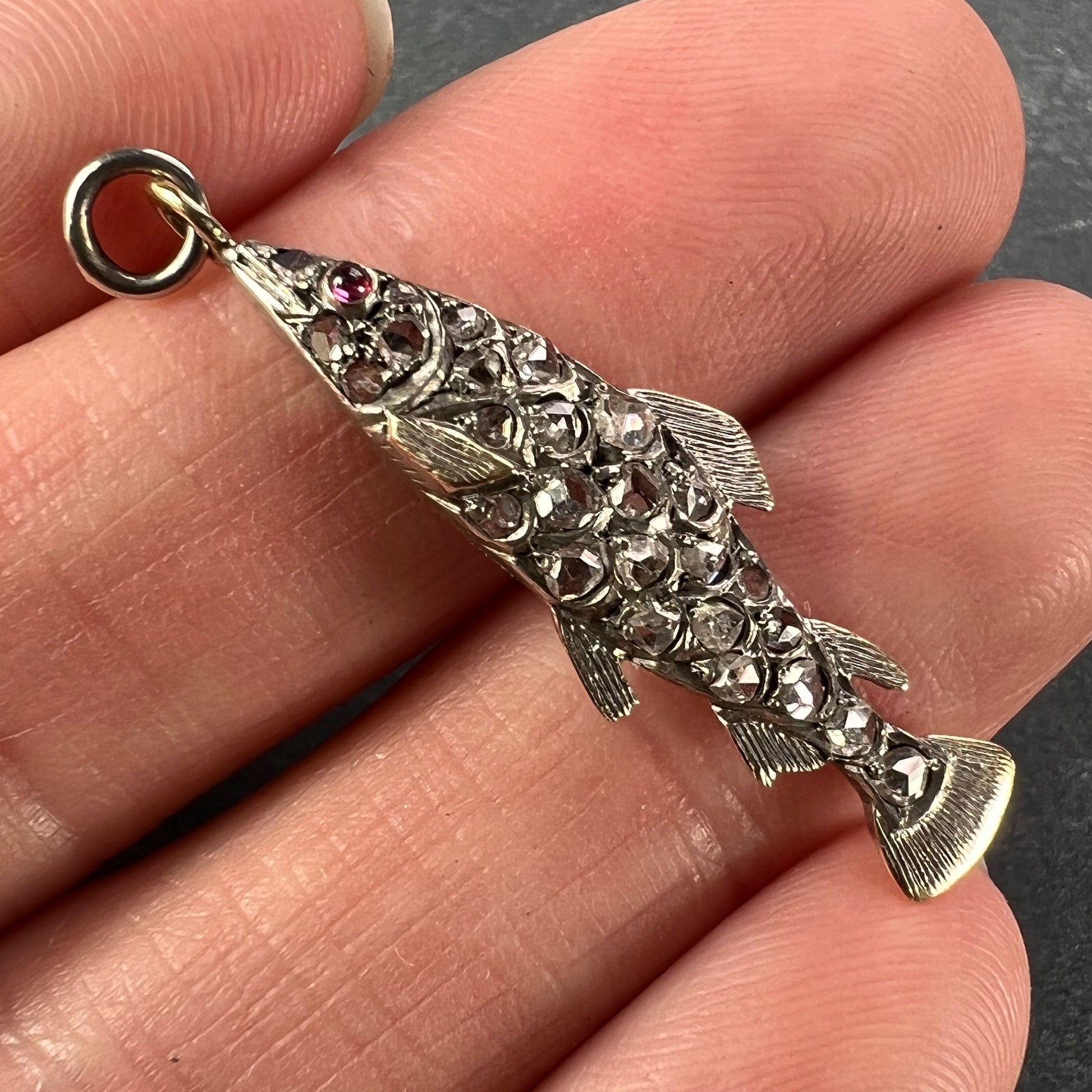 A fish shaped pendant with rose cut diamonds and ruby eye, held in a hand on a grey background