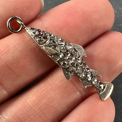 A fish shaped pendant with rose cut diamonds and ruby eye, held in a hand on a grey background