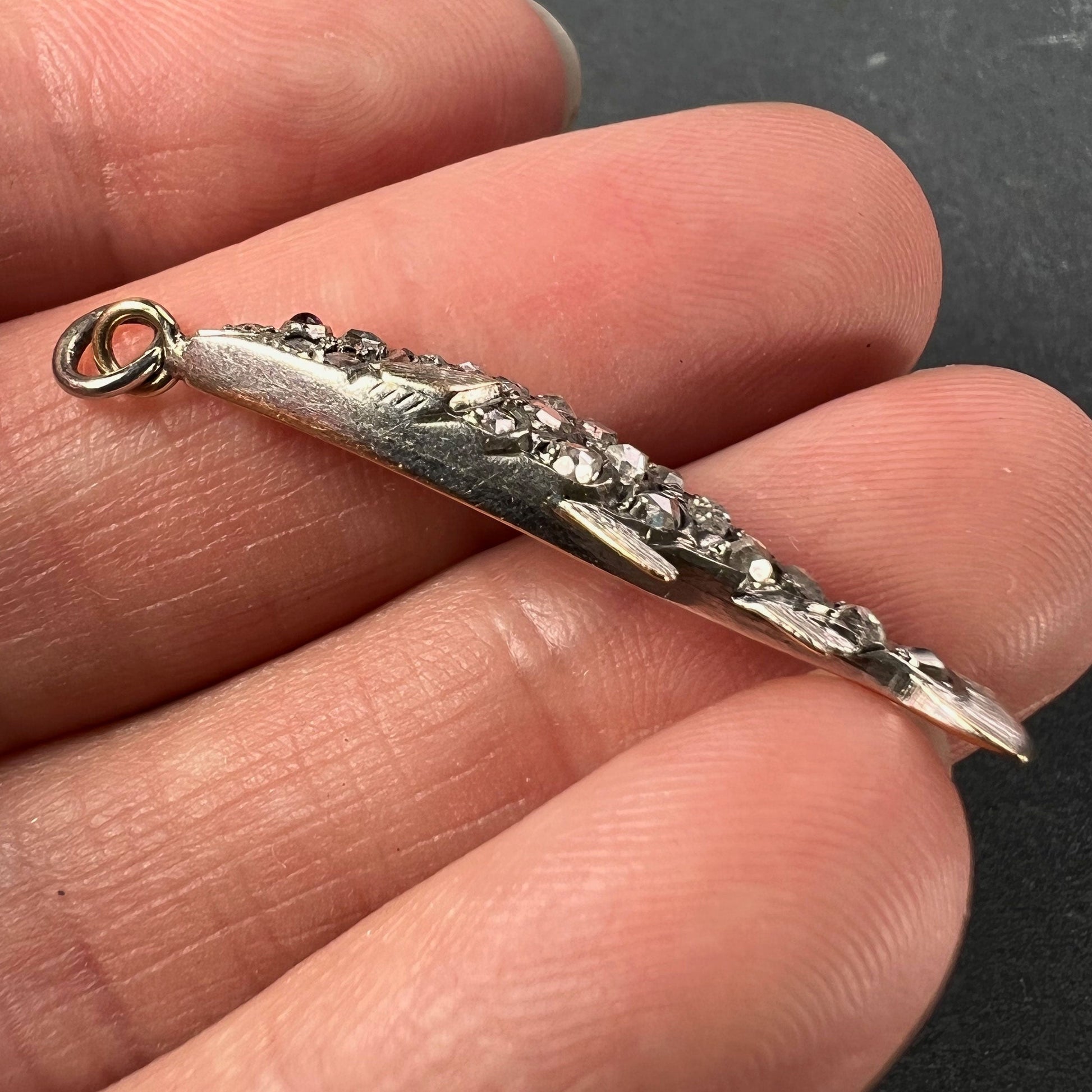 A fish shaped pendant with rose cut diamonds and ruby eye, held in a hand on a grey background