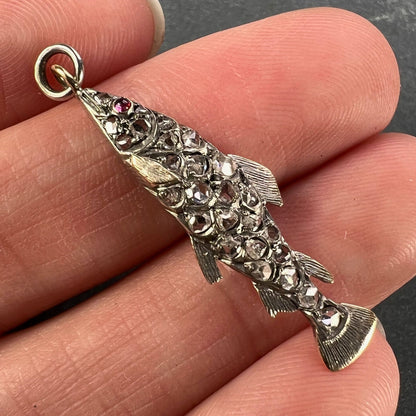 A fish shaped pendant with rose cut diamonds and ruby eye, held in a hand on a grey background