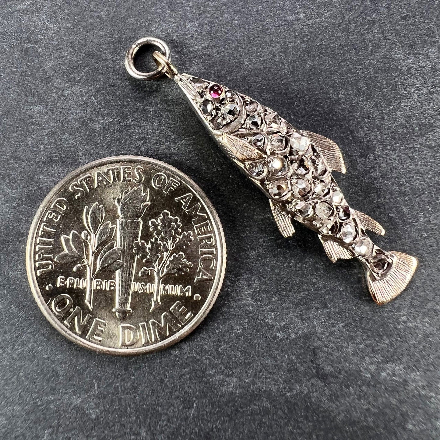 A fish shaped pendant with rose cut diamonds and ruby eye, next to a dime coin on a grey background
