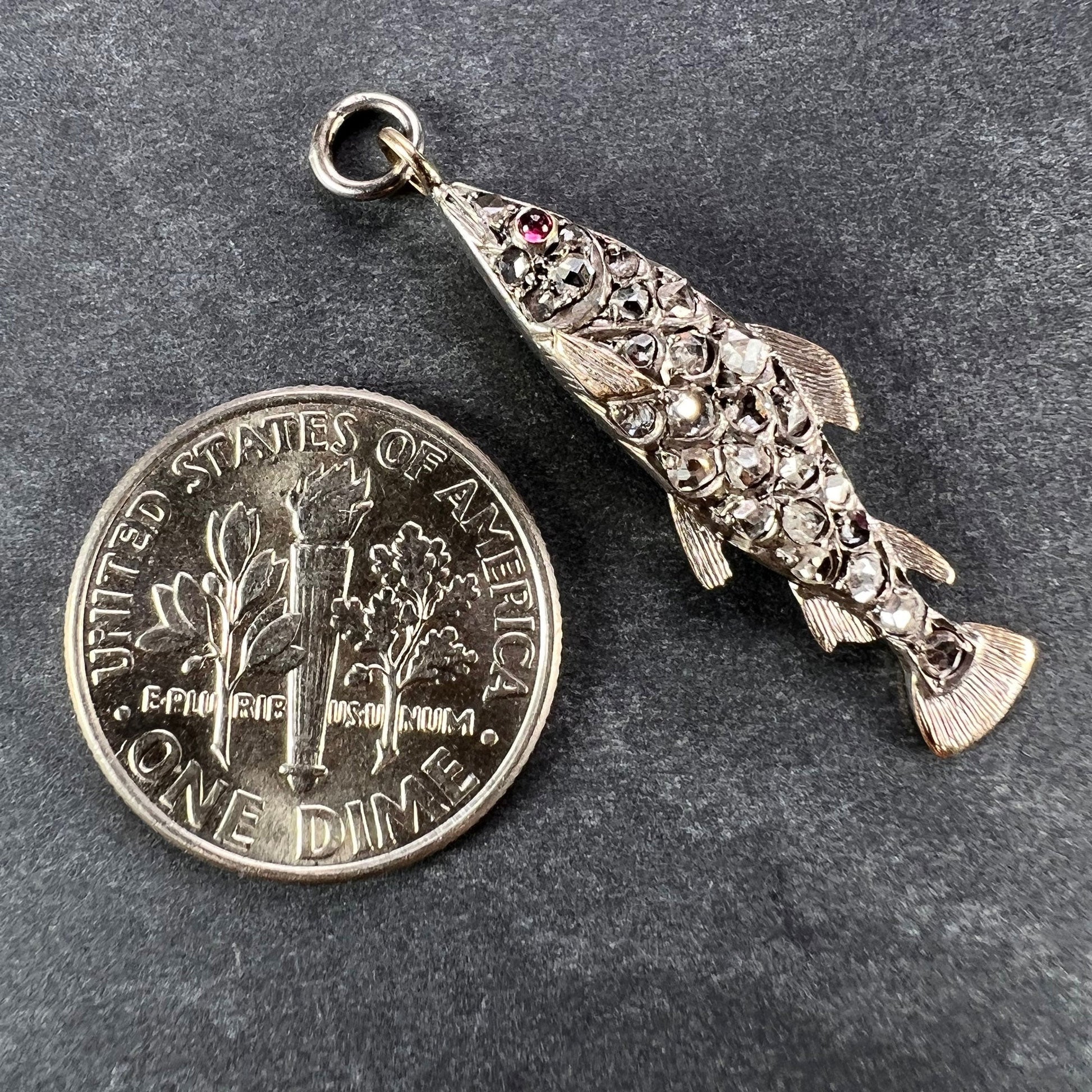 A fish shaped pendant with rose cut diamonds and ruby eye, next to a dime coin on a grey background