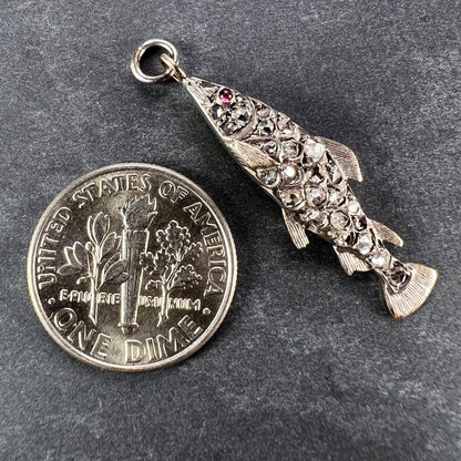 A fish shaped pendant with rose cut diamonds and ruby eye, next to a dime coin on a grey background