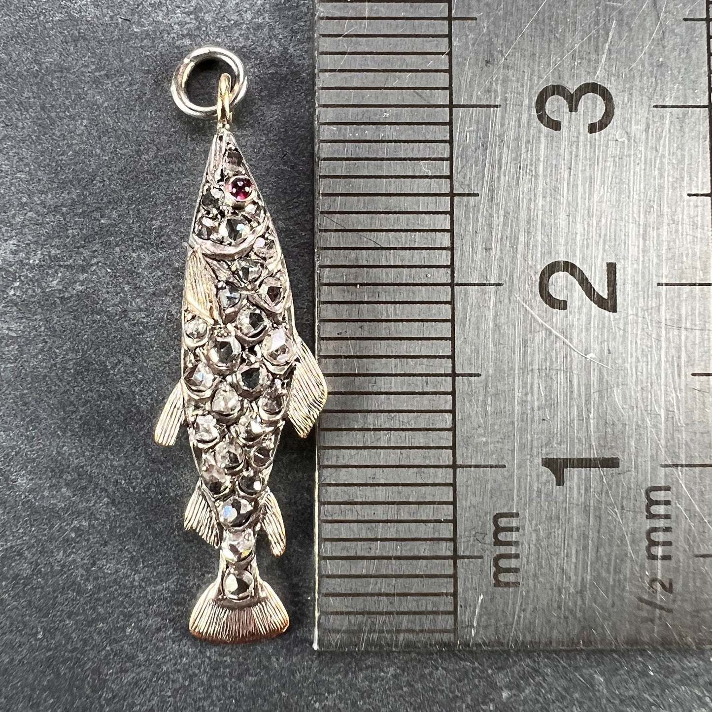 A fish shaped pendant with rose cut diamonds and ruby eye, next to a metal ruler on a grey background