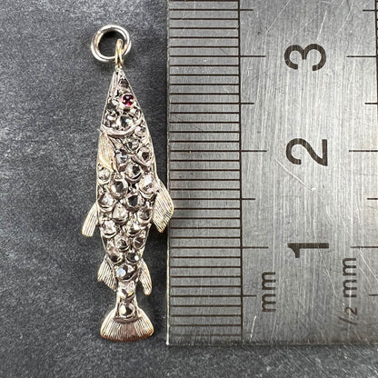 A fish shaped pendant with rose cut diamonds and ruby eye, next to a metal ruler on a grey background