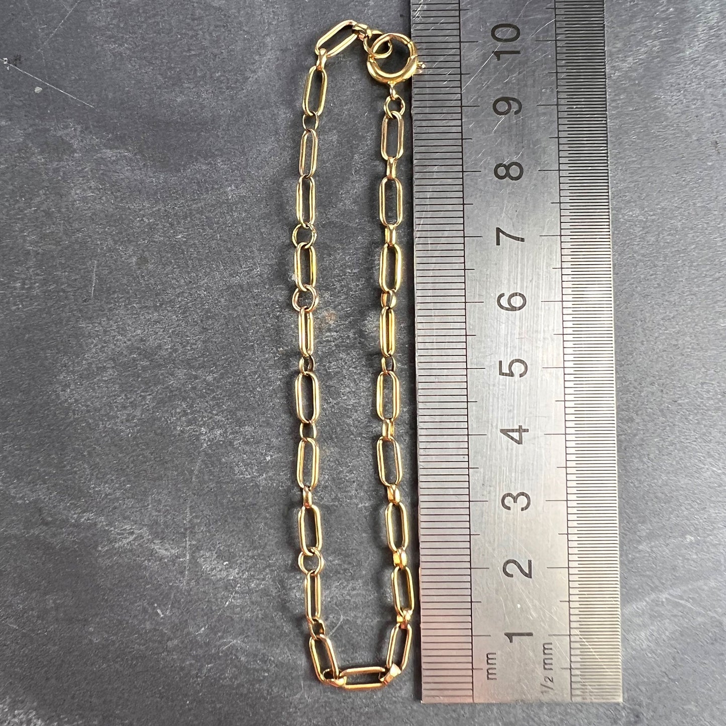 A yellow gold link bracelet with bolt ring clasp, next to a metal ruler on a grey background