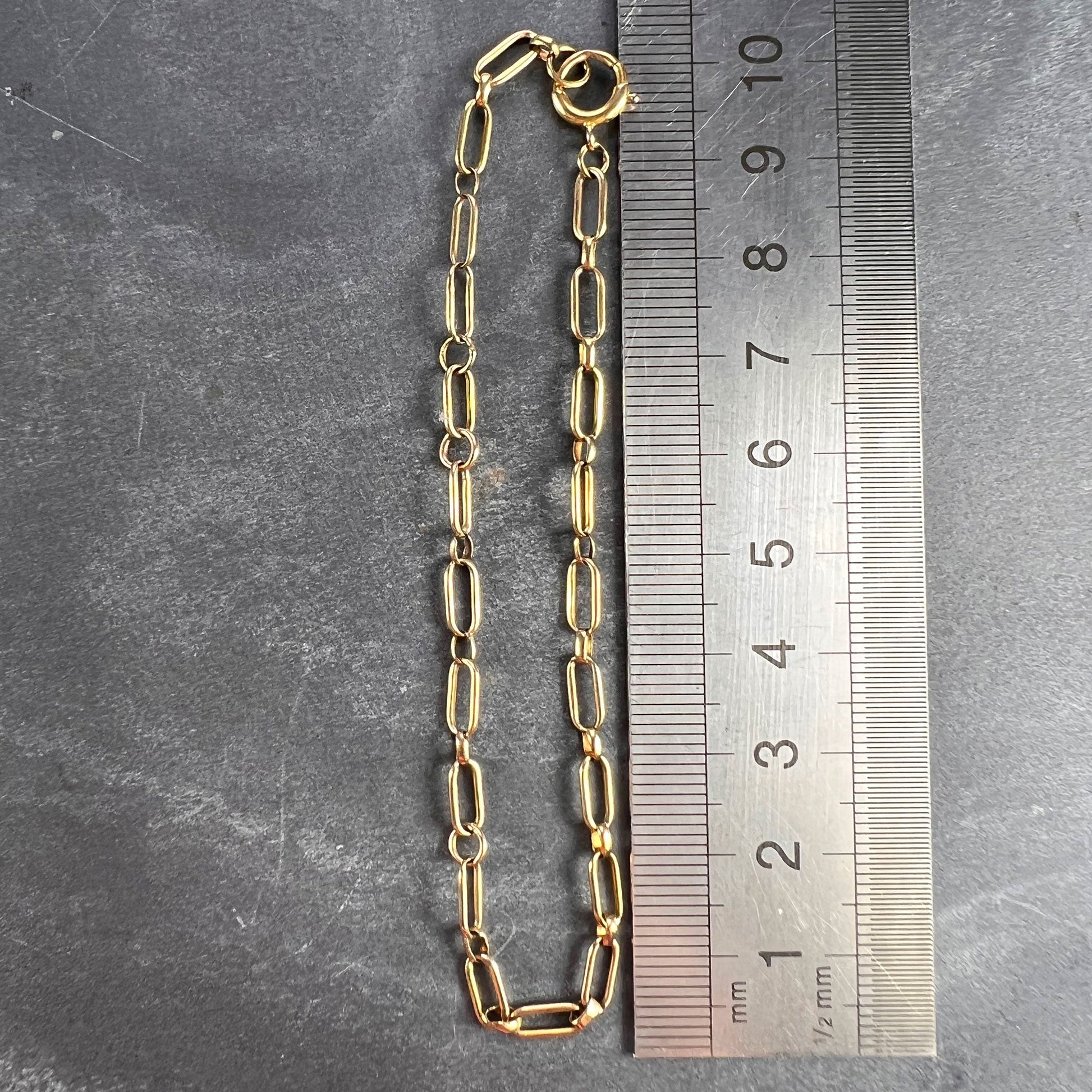 A yellow gold link bracelet with bolt ring clasp, next to a metal ruler on a grey background