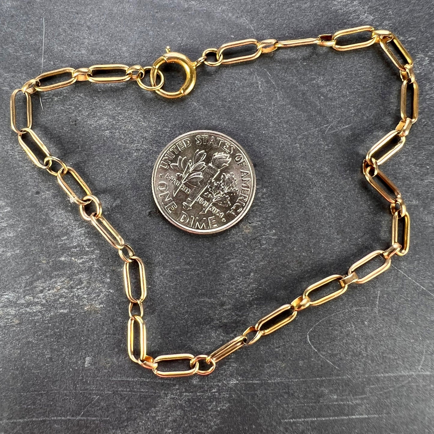 A yellow gold link bracelet with bolt ring clasp, next to a dime coin on a grey background