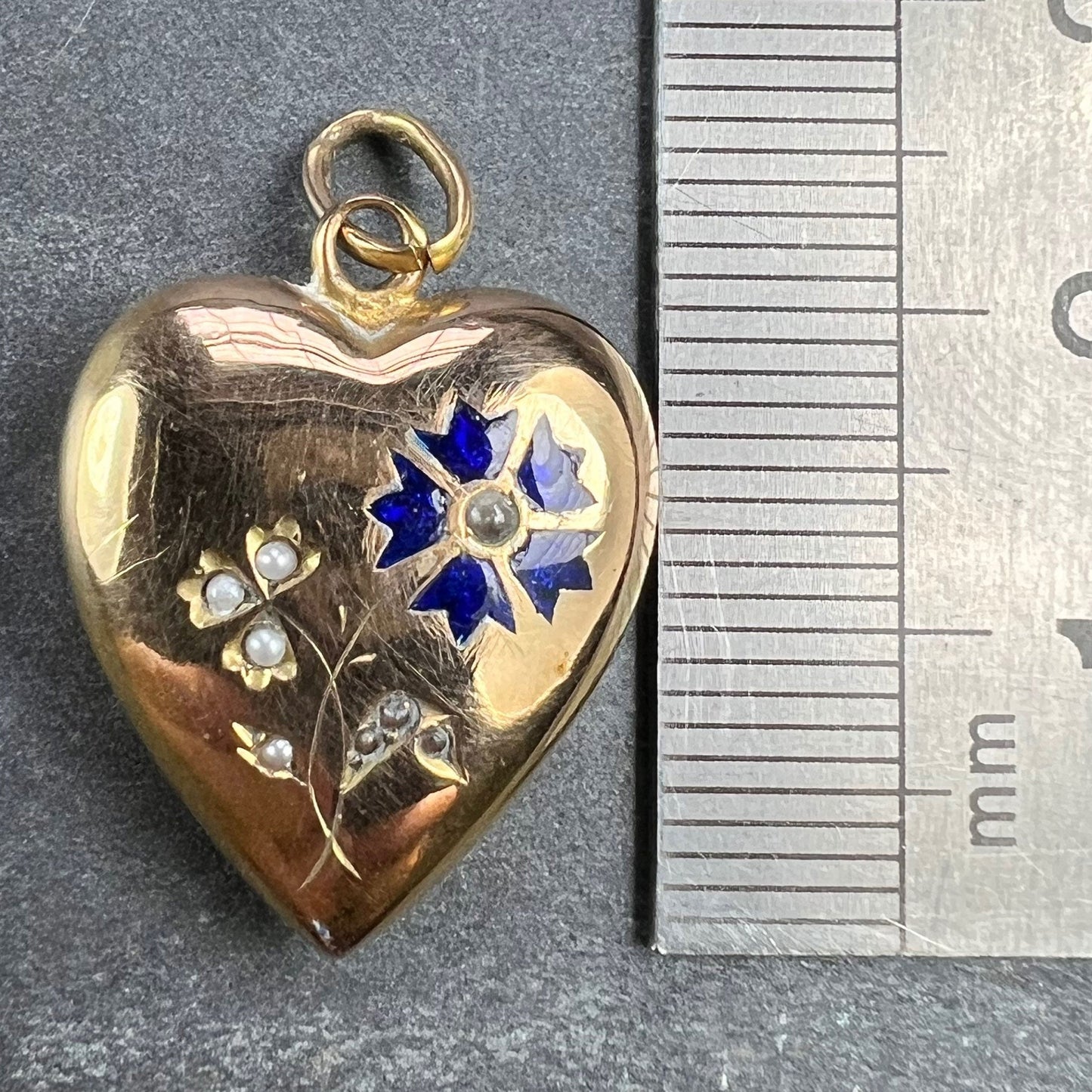 A yellow gold heart shaped pendant with enamel and seed pearls, next to a metal ruler on a grey background
