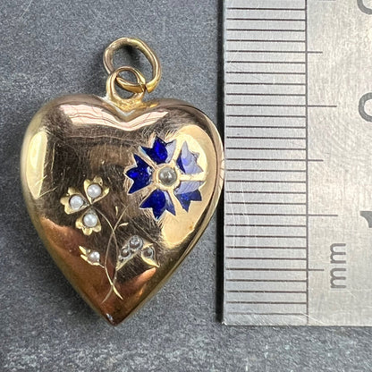 A yellow gold heart shaped pendant with enamel and seed pearls, next to a metal ruler on a grey background