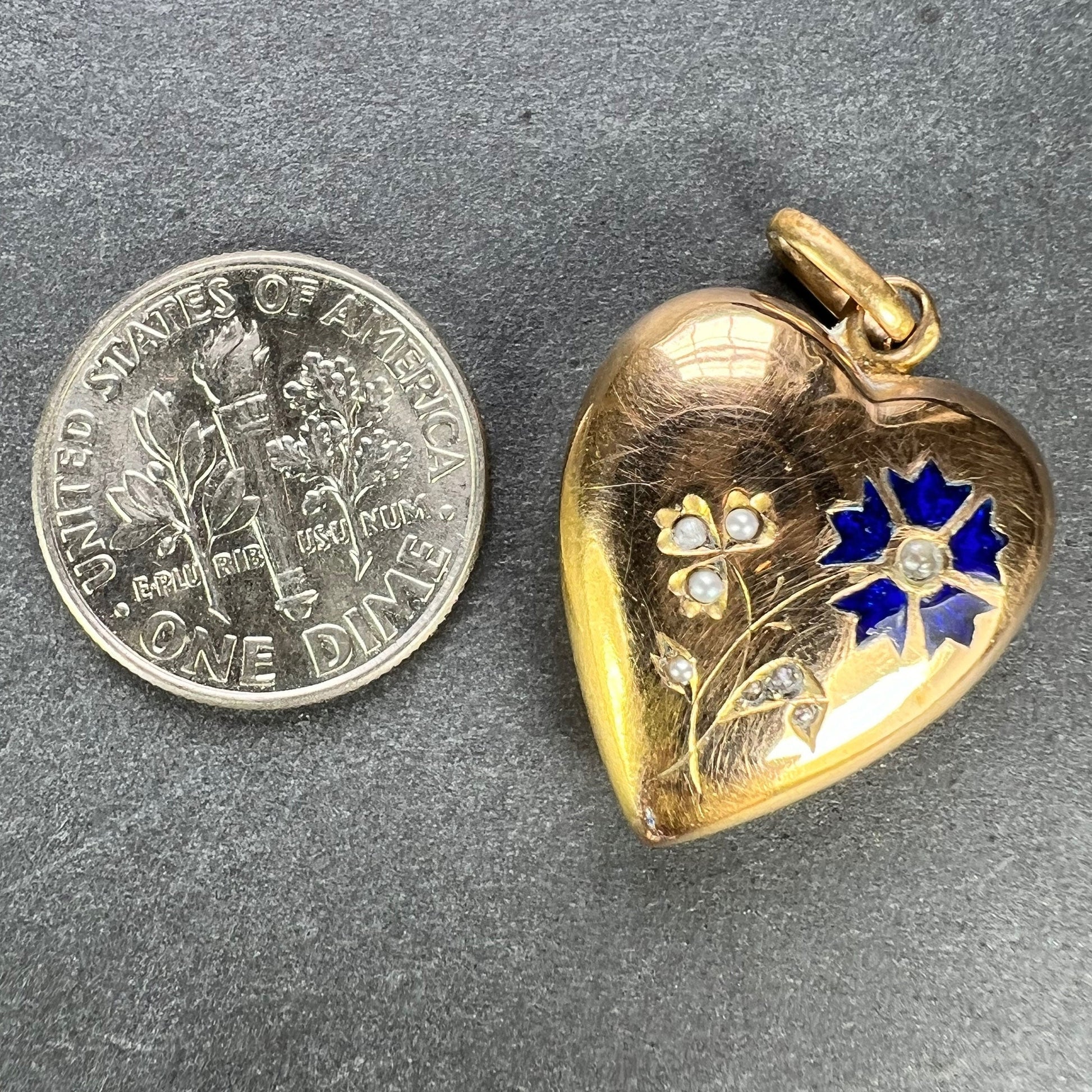 A yellow gold heart shaped pendant with enamel and seed pearls, next to a dime coin on a grey background