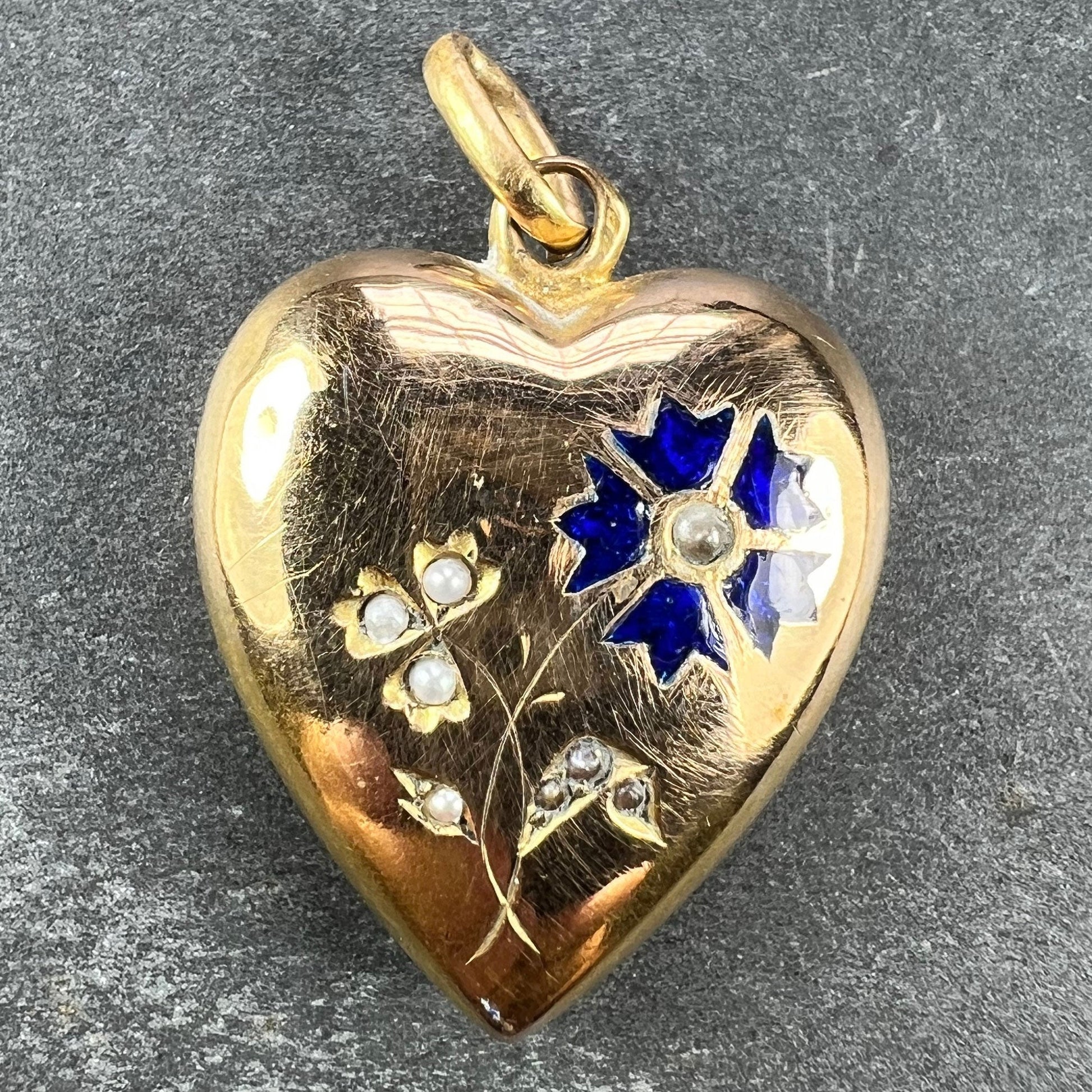 A yellow gold heart shaped pendant with enamel and seed pearls, on a grey background