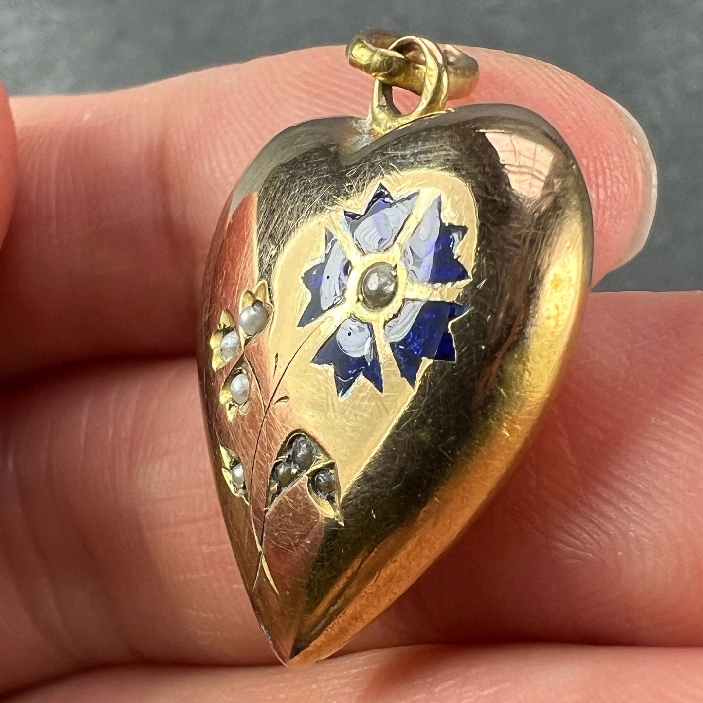 A yellow gold heart shaped pendant with enamel and seed pearls, held in a hand on a grey background