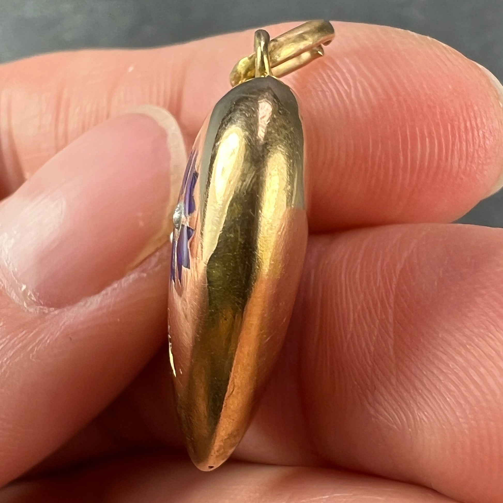 A yellow gold heart shaped pendant with enamel and seed pearls, held in a hand on a grey background