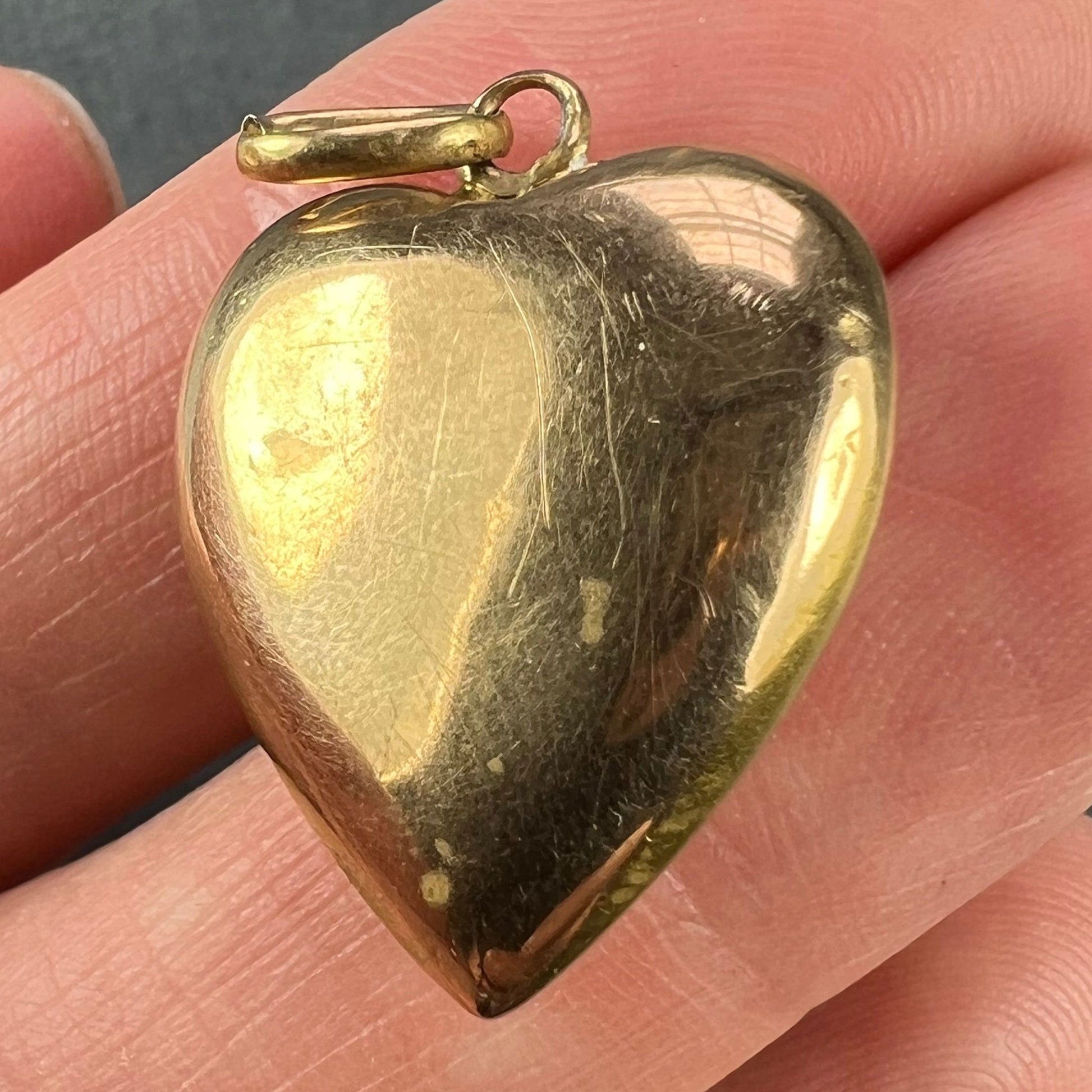 A yellow gold heart shaped pendant, held in a hand on a grey background