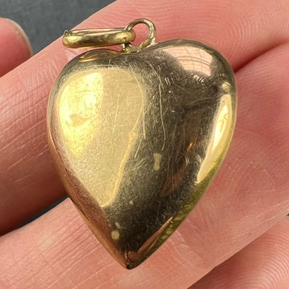 A yellow gold heart shaped pendant, held in a hand on a grey background