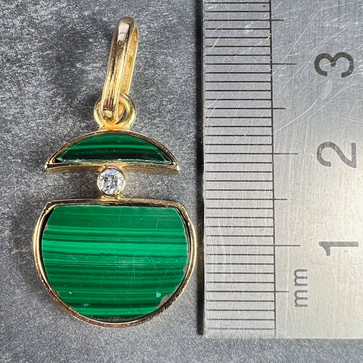 A malachite and diamond yellow gold split circle pendant, next to a metal ruler on a grey background