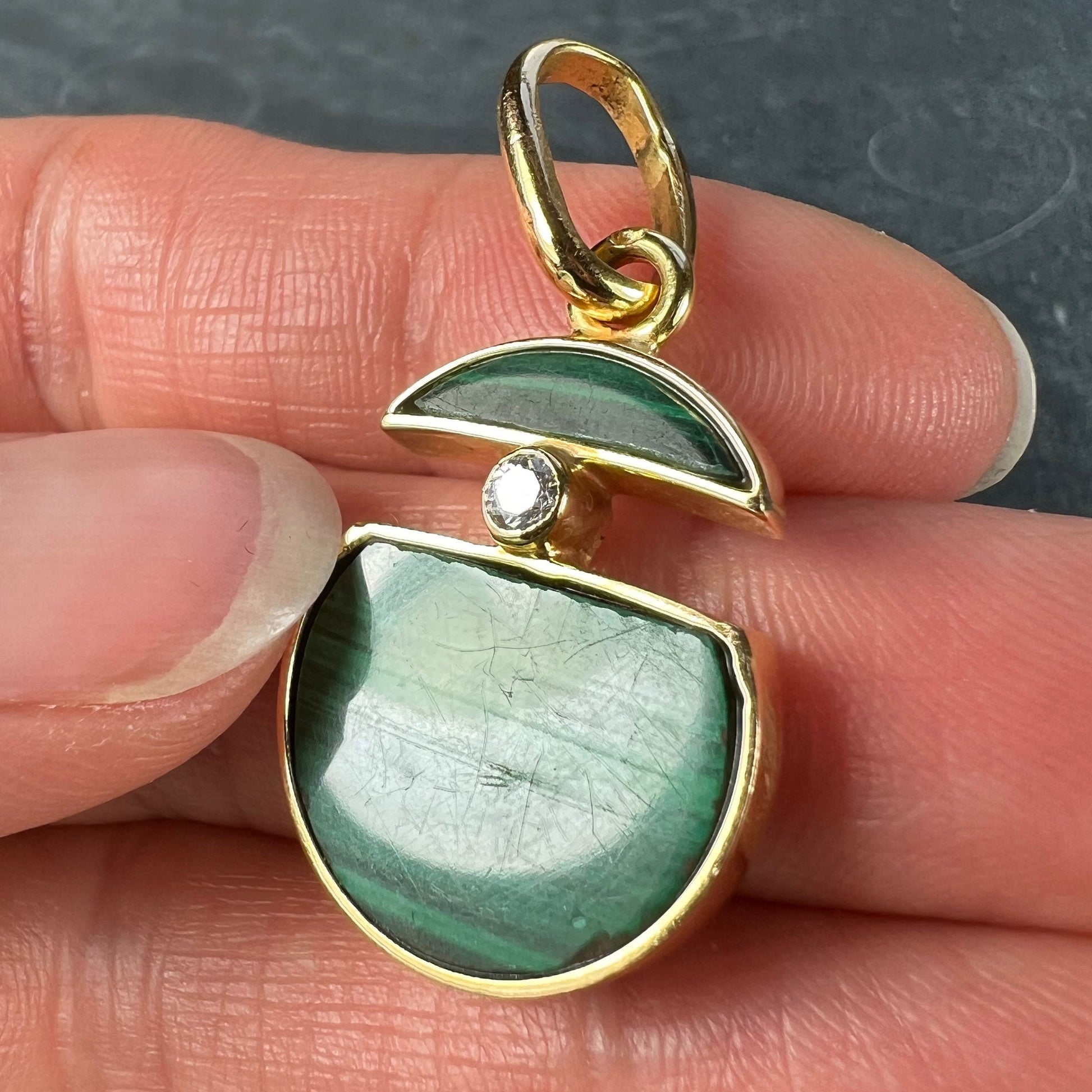 A malachite and diamond yellow gold split circle pendant, held in a hand on a grey background