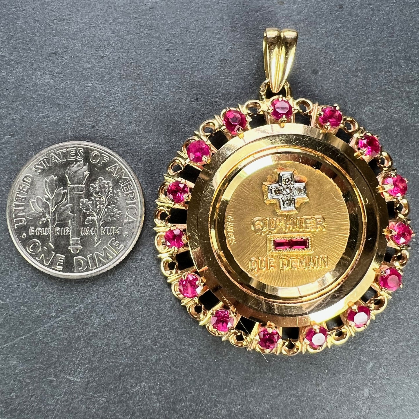 A French QU'HIER QUE DEMAIN pendant with diamonds and synthetic rubies, next to a dime coin on a grey background