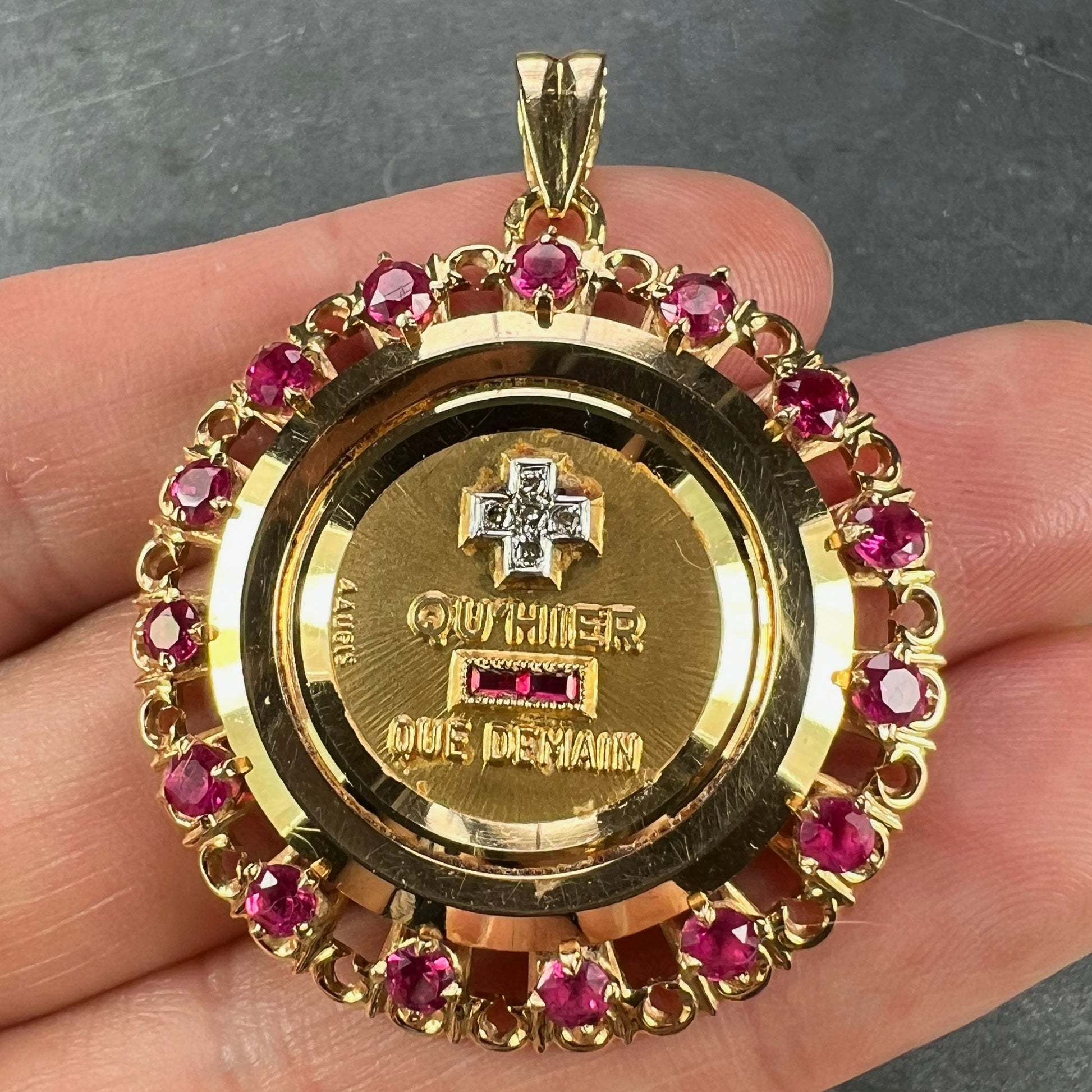 A French QU'HIER QUE DEMAIN pendant with diamonds and synthetic rubies, held in a hand on a grey background