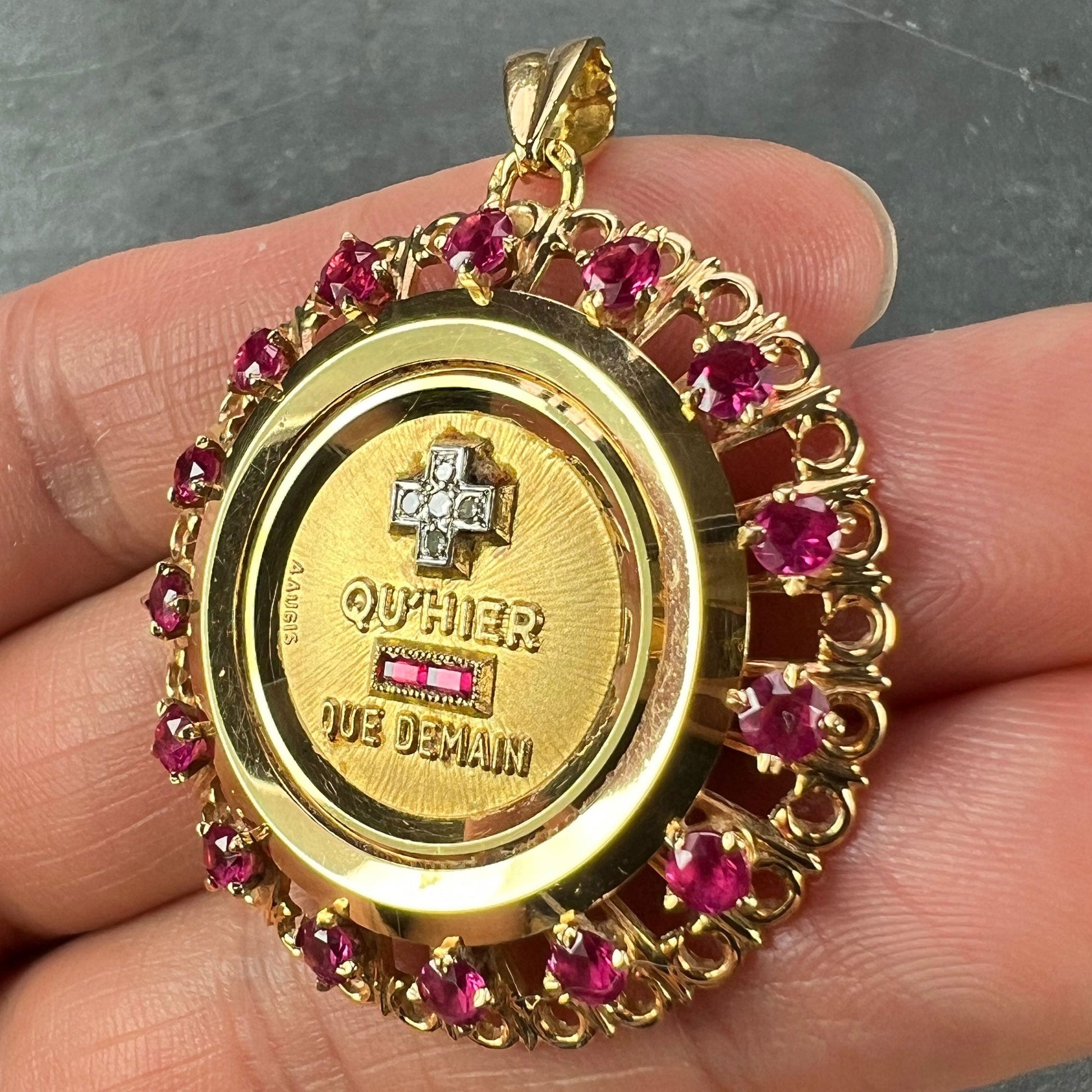 A French QU'HIER QUE DEMAIN pendant with diamonds and synthetic rubies, held in a hand on a grey background