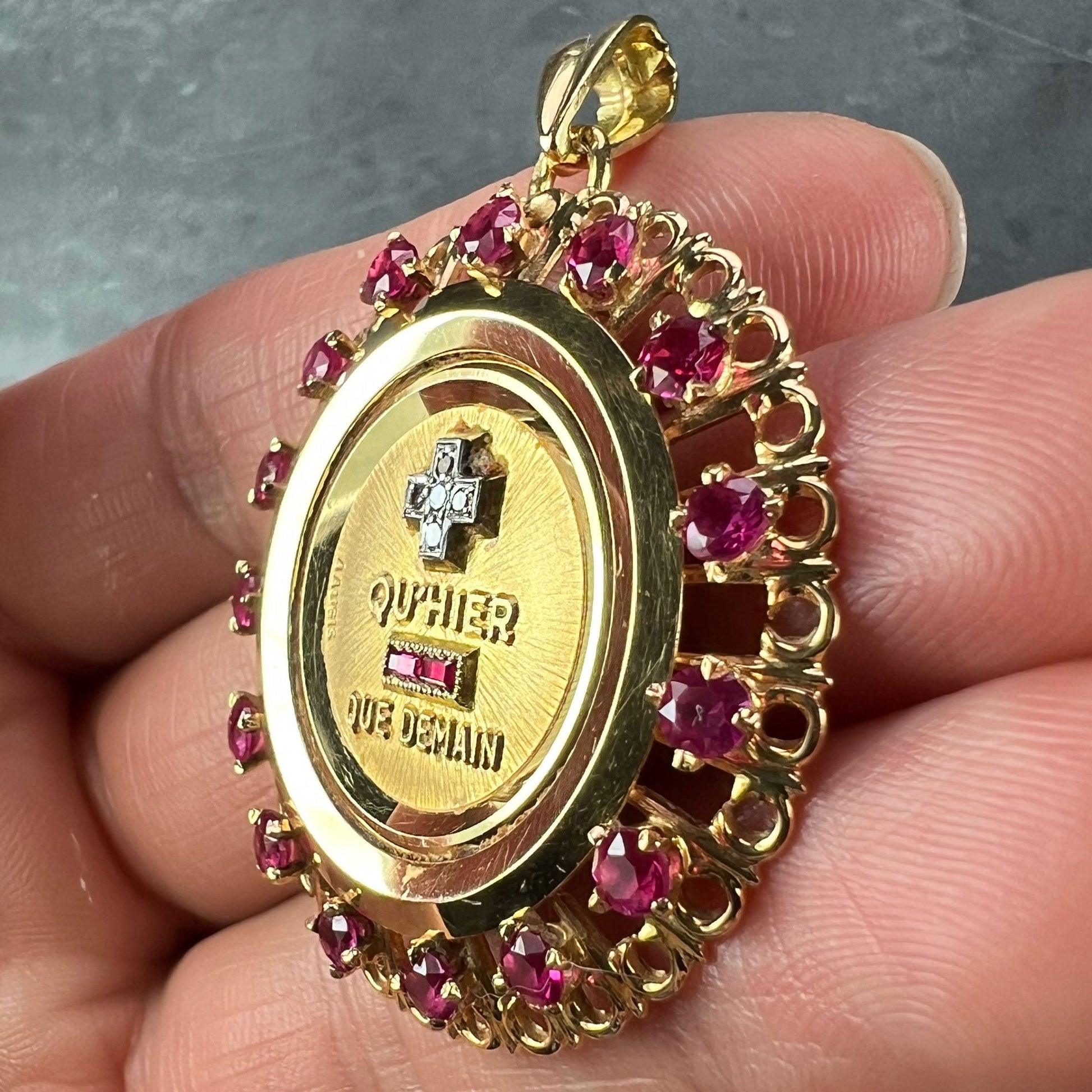 A French QU'HIER QUE DEMAIN pendant with diamonds and synthetic rubies, held in a hand on a grey background