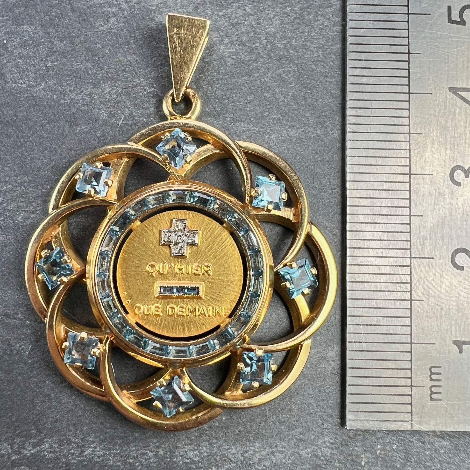 A yellow gold French QU'HIER QUE DEMAIN pendant with blue stones and diamonds, next to a metal ruler on a grey background