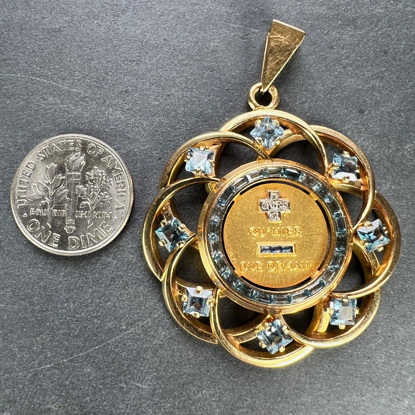 A yellow gold French QU'HIER QUE DEMAIN pendant with blue stones and diamonds, next to a dime coin on a grey background