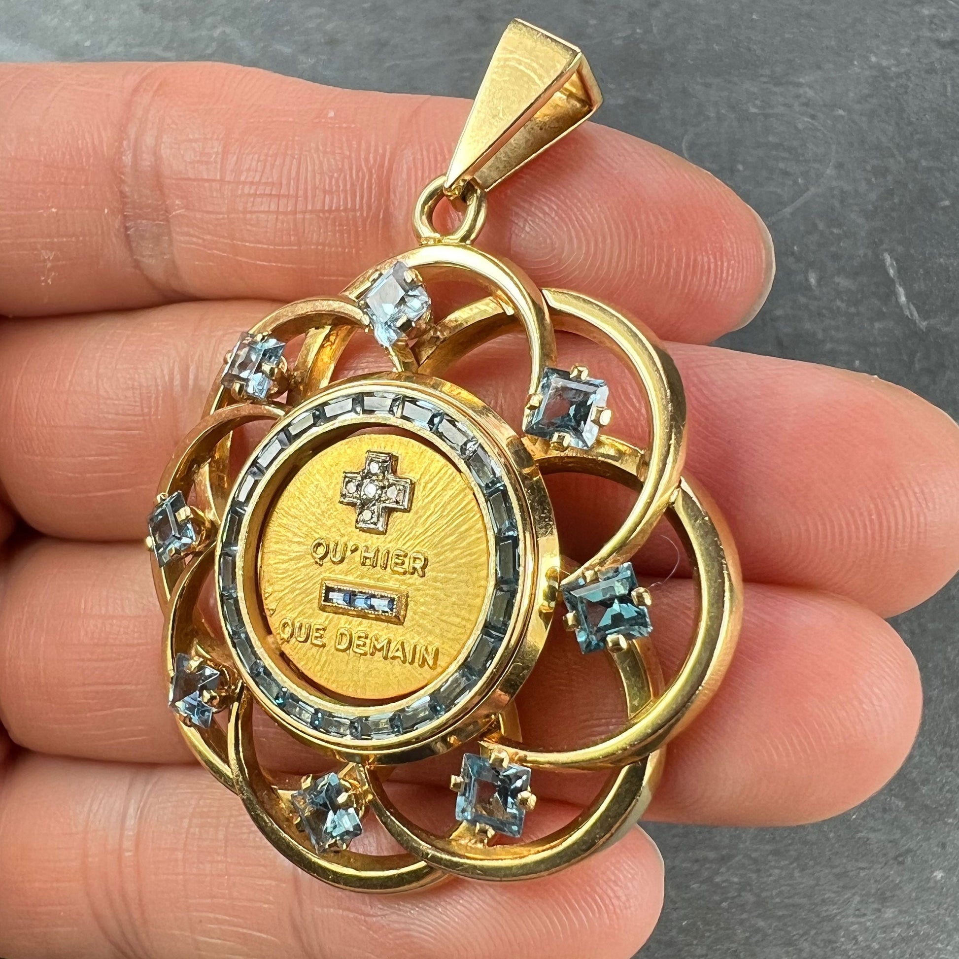 A yellow gold French QU'HIER QUE DEMAIN pendant with blue stones and diamonds, held in a hand on a grey background