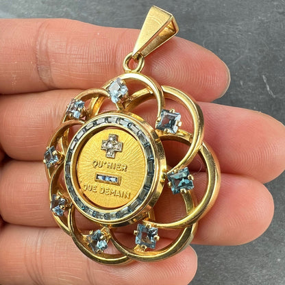 A yellow gold French QU'HIER QUE DEMAIN pendant with blue stones and diamonds, held in a hand on a grey background