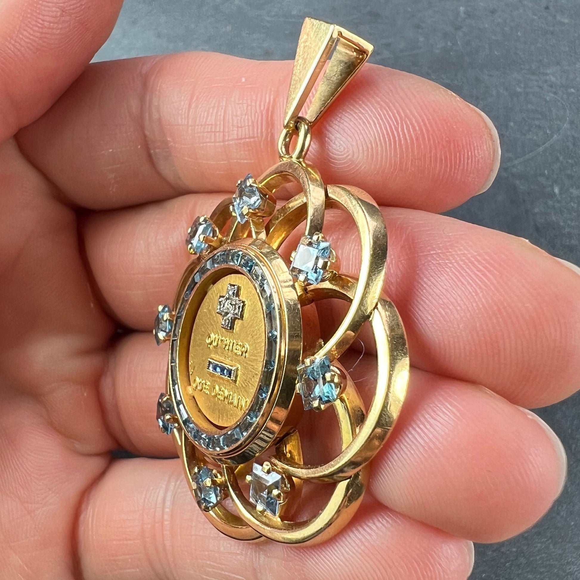 A yellow gold French QU'HIER QUE DEMAIN pendant with blue stones and diamonds, held in a hand on a grey background